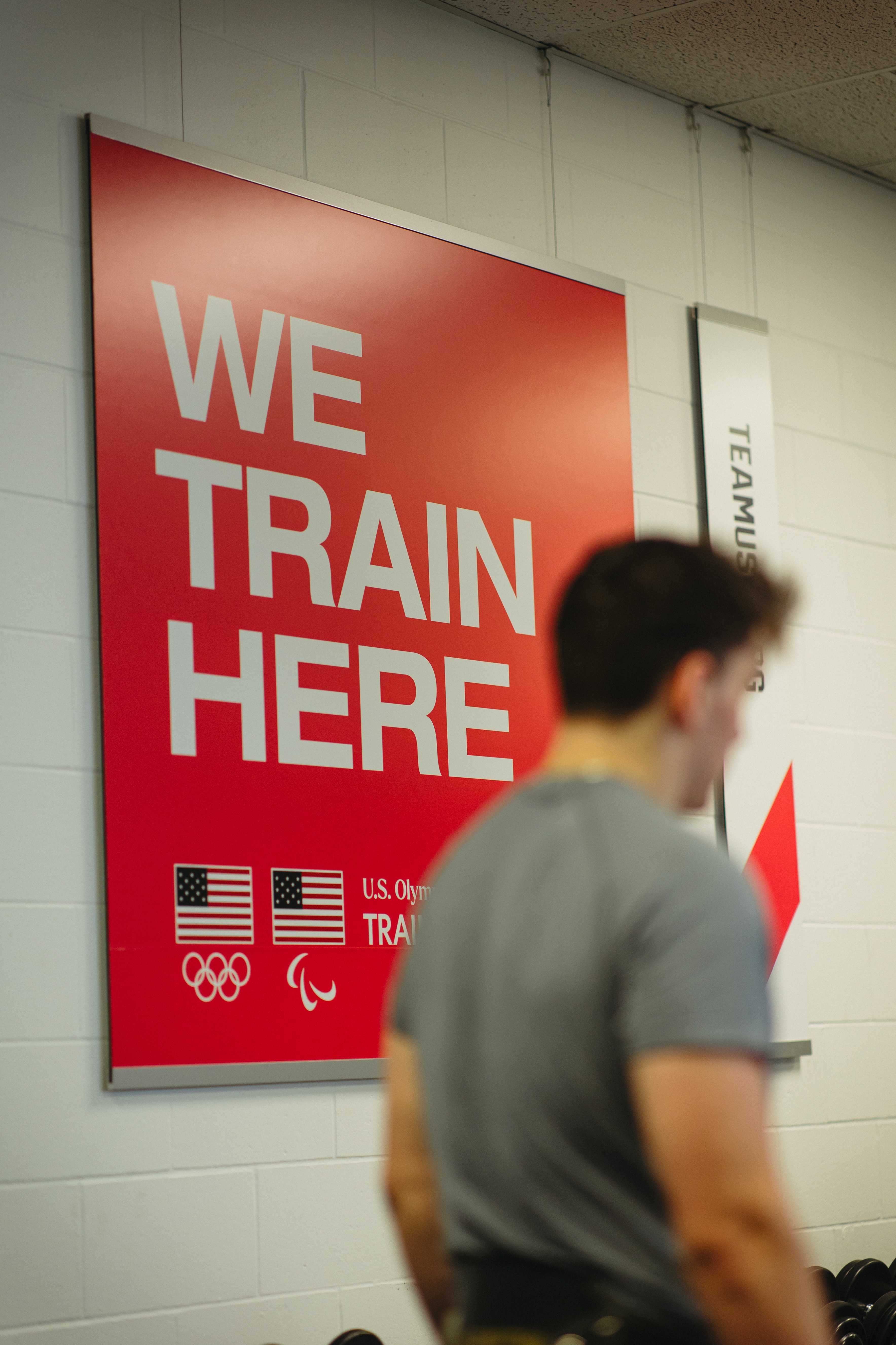 Logan Barnes training in the gym beneath a red 'We Train Here' U.S. Olympic & Paralympic Training Center sign