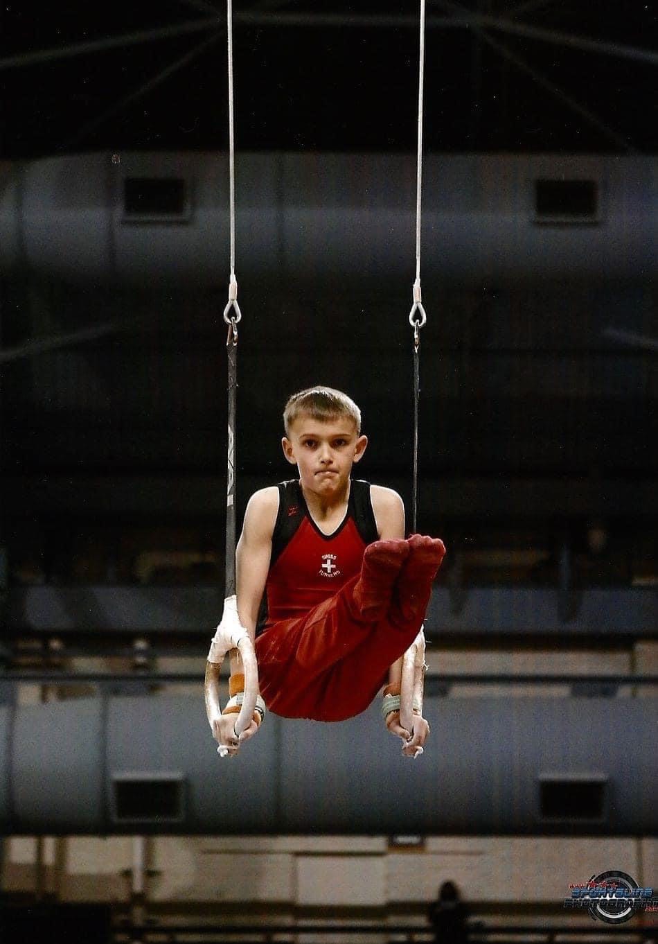 Young Logan Barnes performing on gymnastics rings during competition, showing his athletic foundation before discovering luge